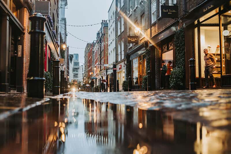 An image of a rainy street lined with shops and streetlights.